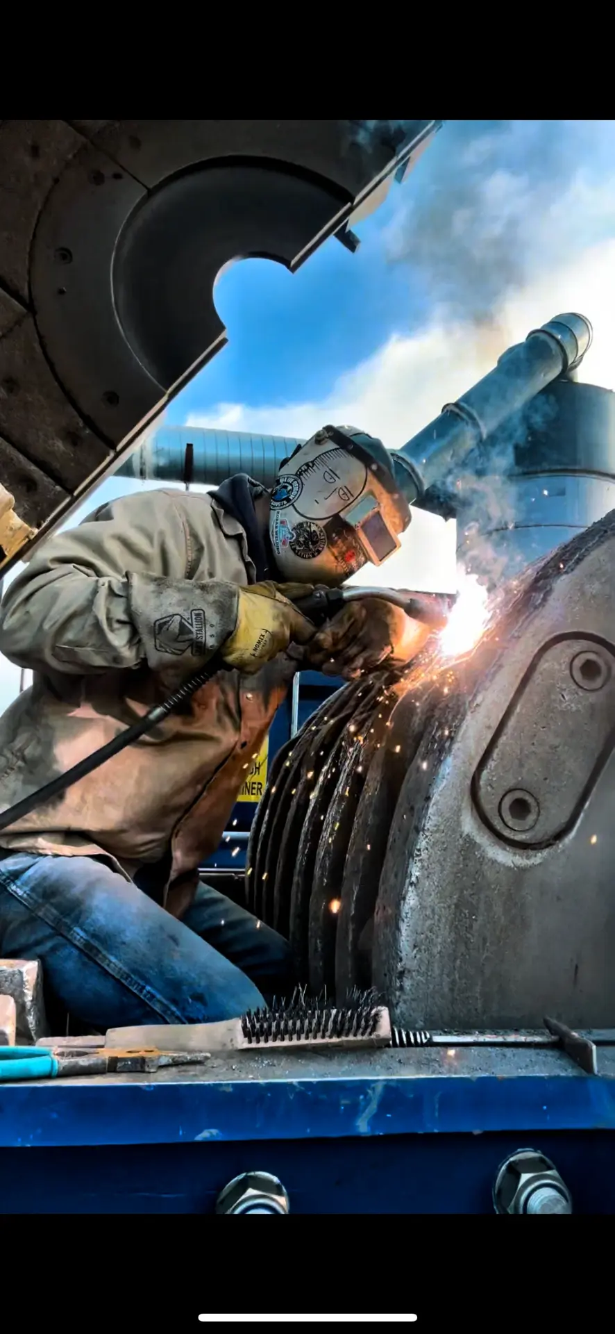 Welder crouched on a heavy steel rotor assembly welding stacked steel discs at an industrial site