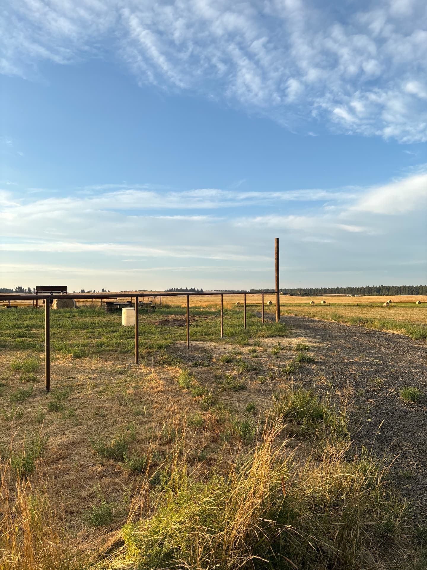 Completed welded steel pipe fence line running along a gravel ranch road with a livestock feeder and hay bales in the pasture