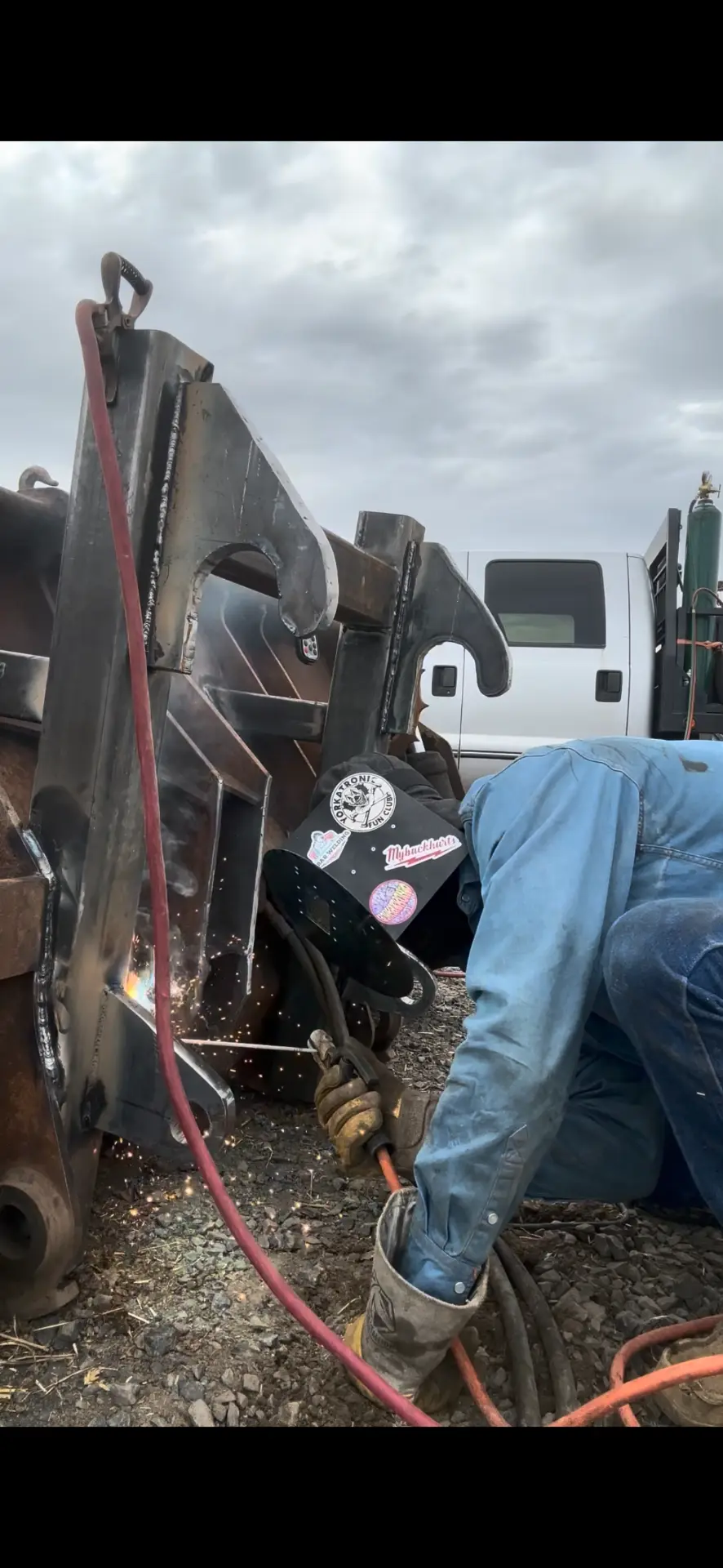 Kirby Orr welding steel brackets onto heavy equipment at a job site, with the Kobar Welding mobile truck and gas cylinder behind